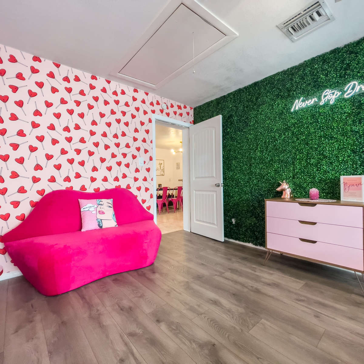 The room features a pink sofa against a wall with heart-patterned wallpaper and a green foliage wall with a neon sign that reads "New Beg Dreaming."