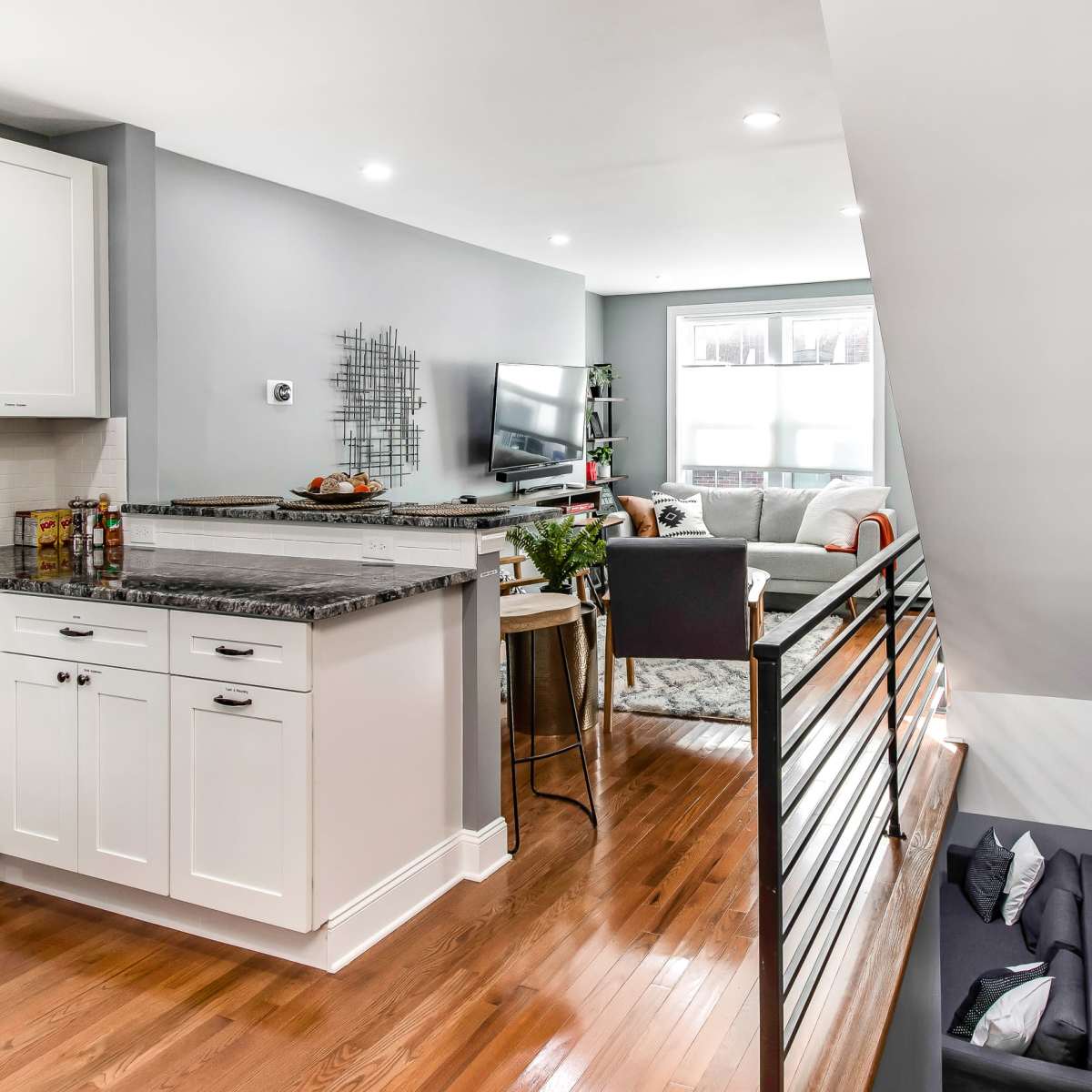 A modern kitchen with white cabinets and granite countertops is seamlessly connected to a living area featuring a sofa and television, all within a well-lit space.
