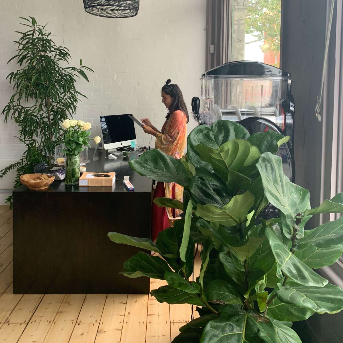 A woman stands at a desk with a computer in a bright, plant-filled room featuring large windows and decorative light fixtures.