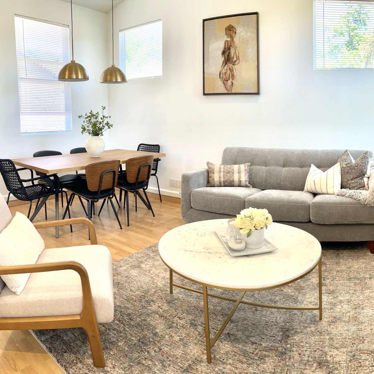 A modern living room with a gray sofa, a round marble coffee table, a wooden side table, and a dining area featuring a table and black chairs in a well-lit space.