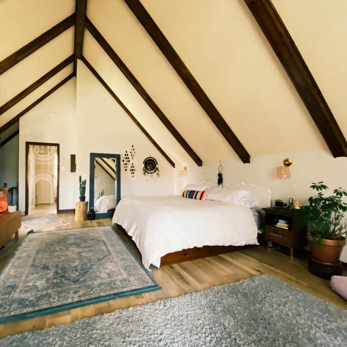 A spacious bedroom with a high, vaulted ceiling, featuring a large bed, a television mounted on the wall, and plants placed around the room.