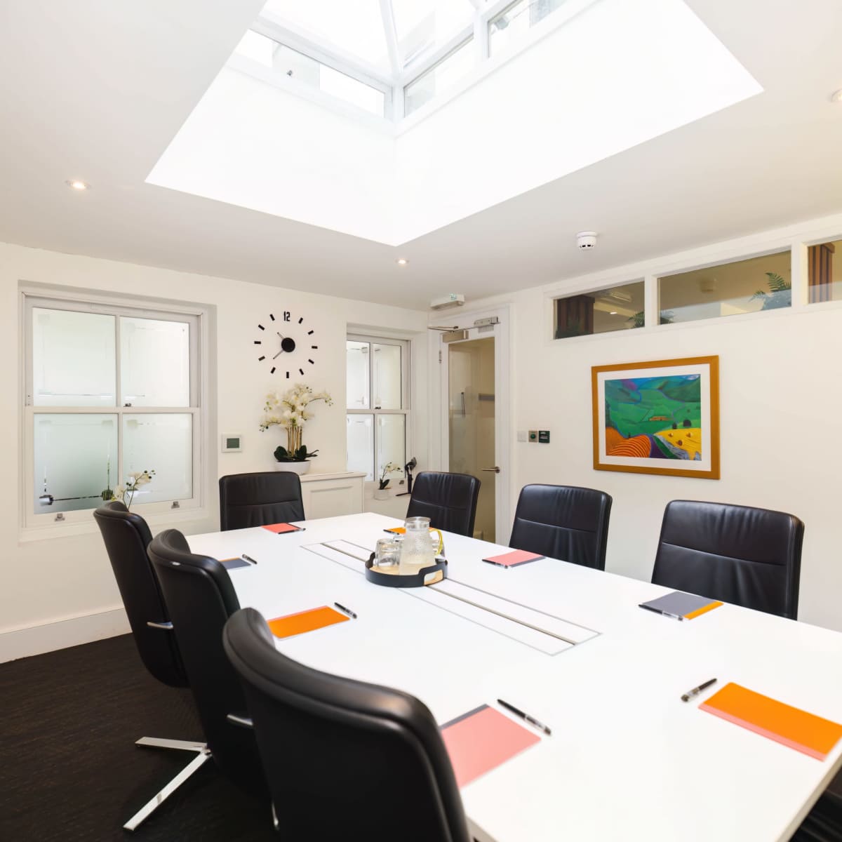 A modern conference room features a white table surrounded by black chairs, with artwork on the walls and a skylight above.