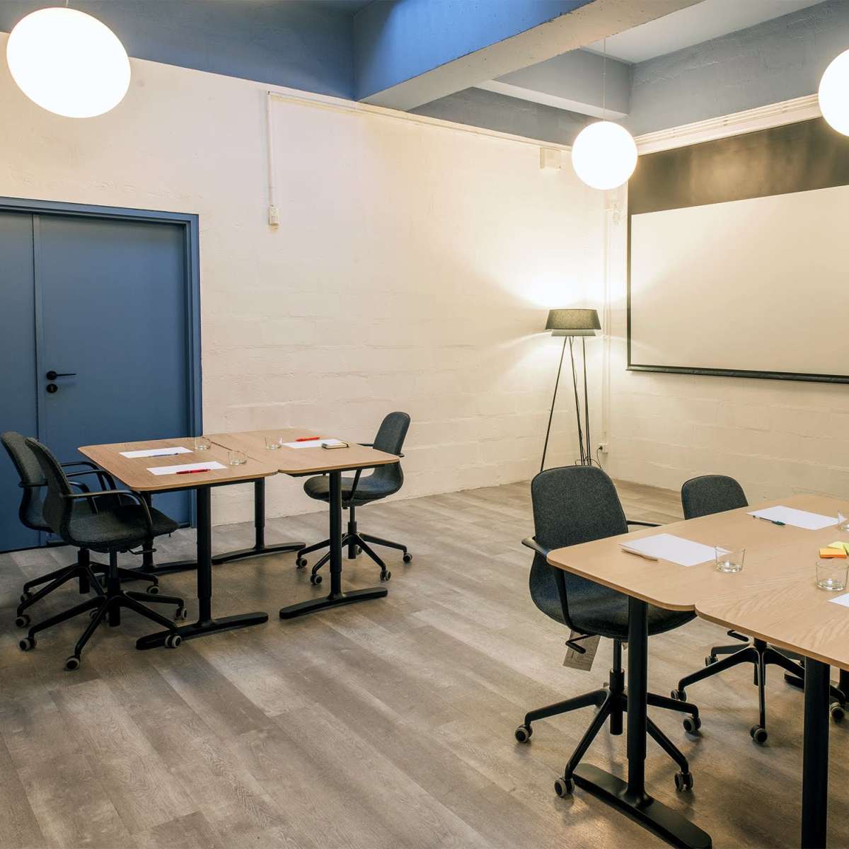 A bright, modern meeting room with two tables arranged for a discussion, each equipped with notepads, pens, and glasses of water, alongside a projector screen at the front.