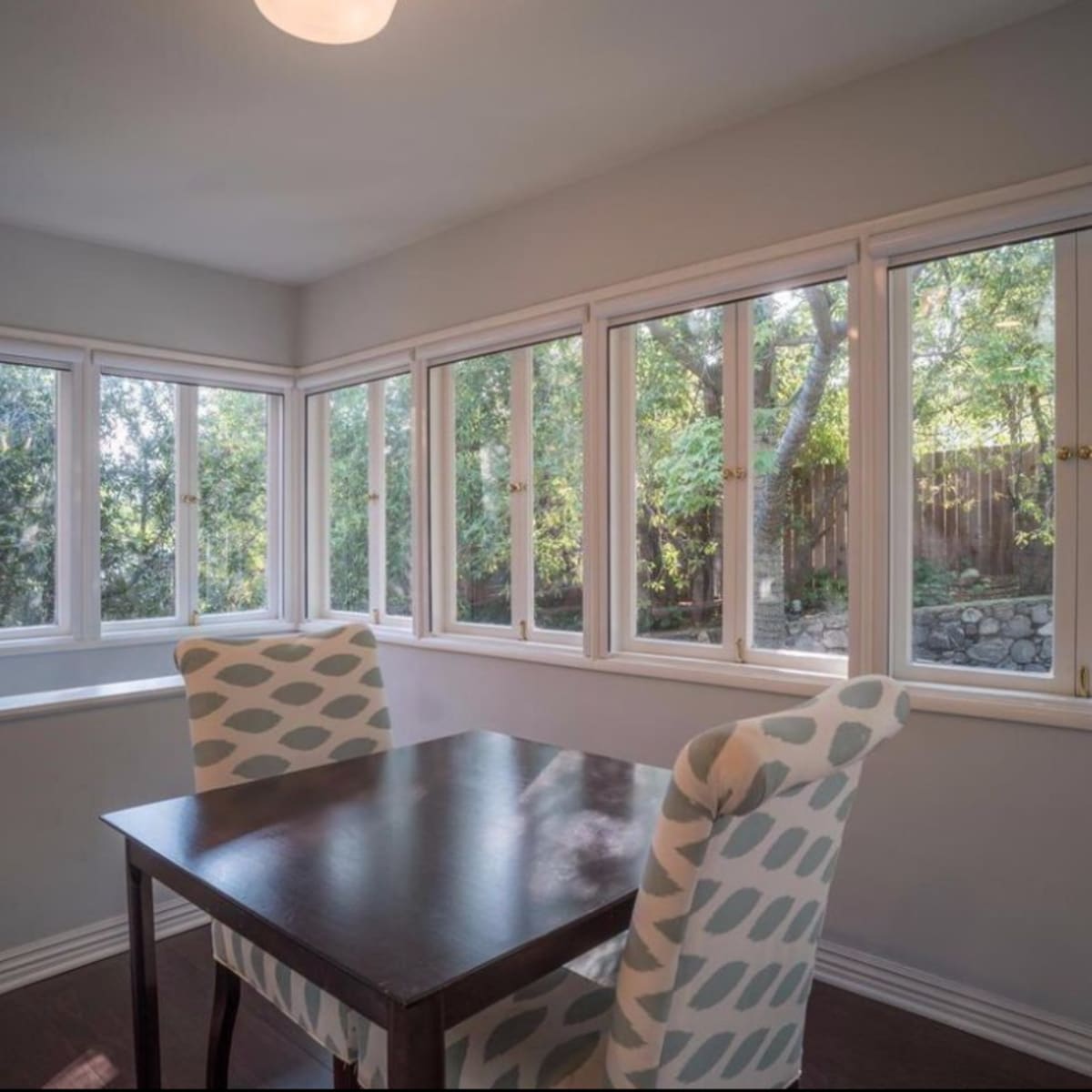 A dining area features a brown table next to two patterned chairs, surrounded by large windows that overlook a garden.