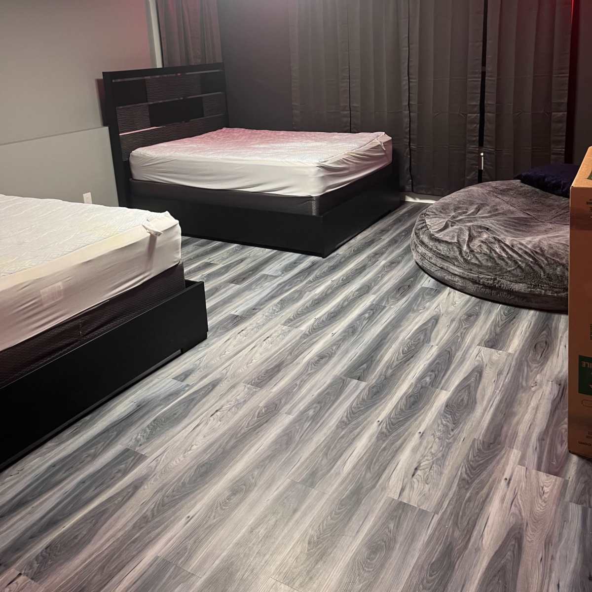 A room with two unmade beds, a round gray beanbag chair, and dark curtains covering the windows.