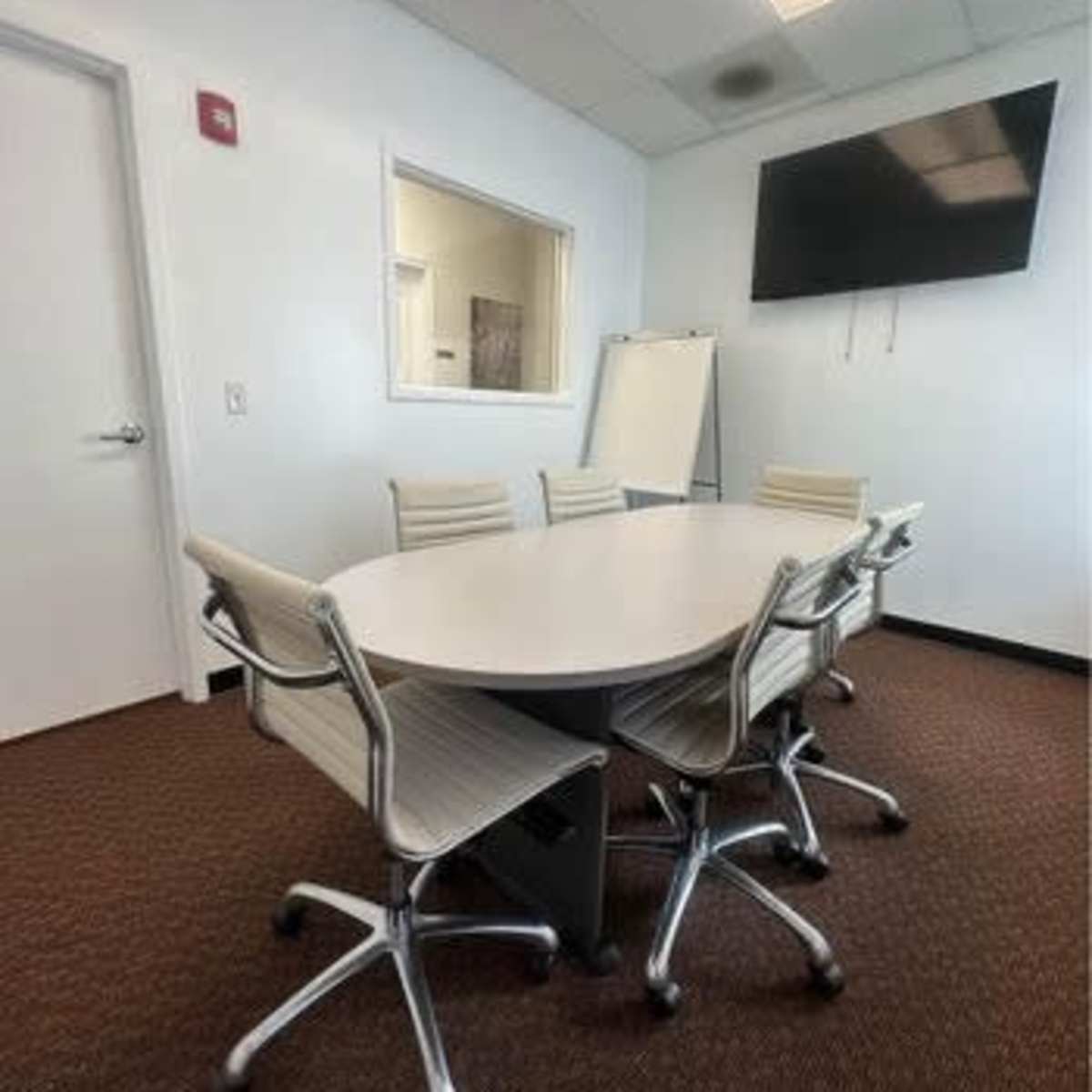 A conference room with a large oval table surrounded by six modern chairs and a wall-mounted television.