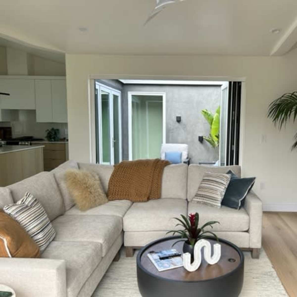 A light-filled living room features a beige sectional sofa, a round coffee table with decorative items, and a potted plant next to an open sliding door leading to an outdoor space.