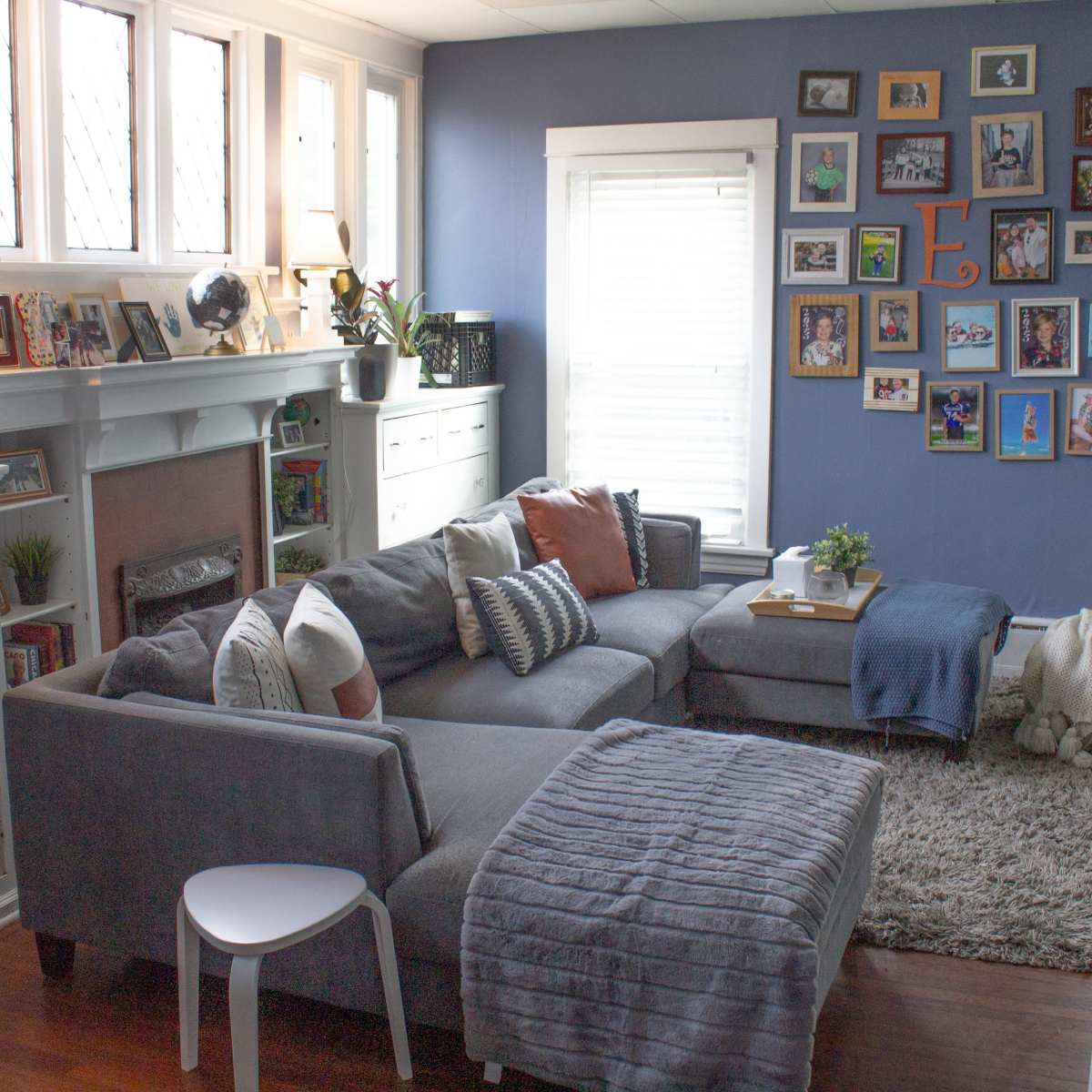 A cozy living room features a gray sectional sofa with patterned pillows, a coffee table on a plush rug, and a gallery wall of framed photos and artwork.