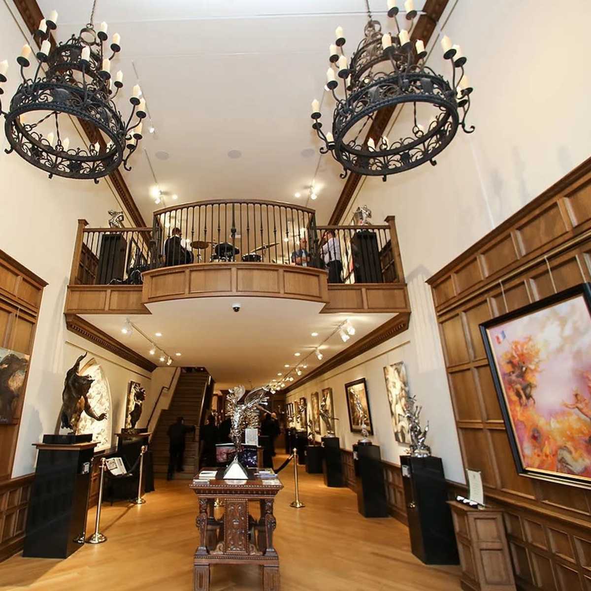 The interior of an art gallery features wooden paneling, multiple large paintings, and a staircase leading to a second level.