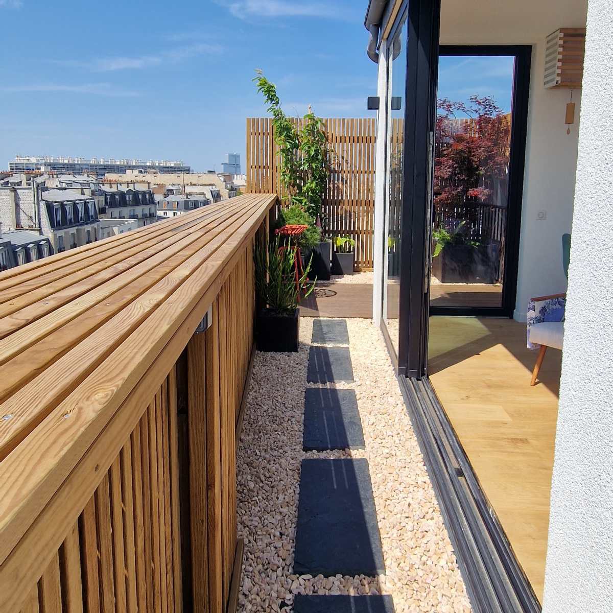 A rooftop terrace with wooden railing, stone pathways, and potted plants under a clear blue sky.