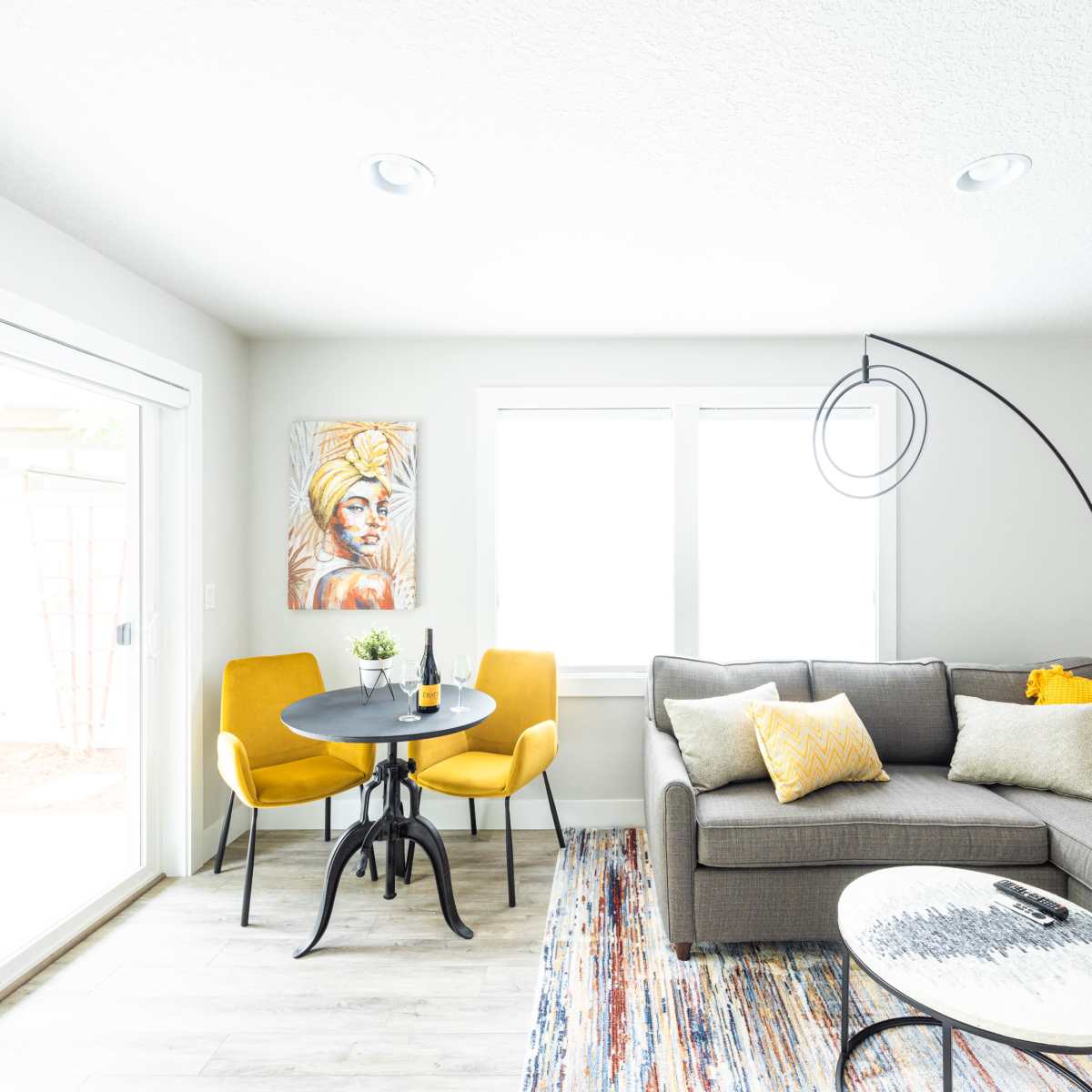 A modern living room with a gray sectional sofa, a circular dining table with two yellow chairs, and large windows that provide natural light.