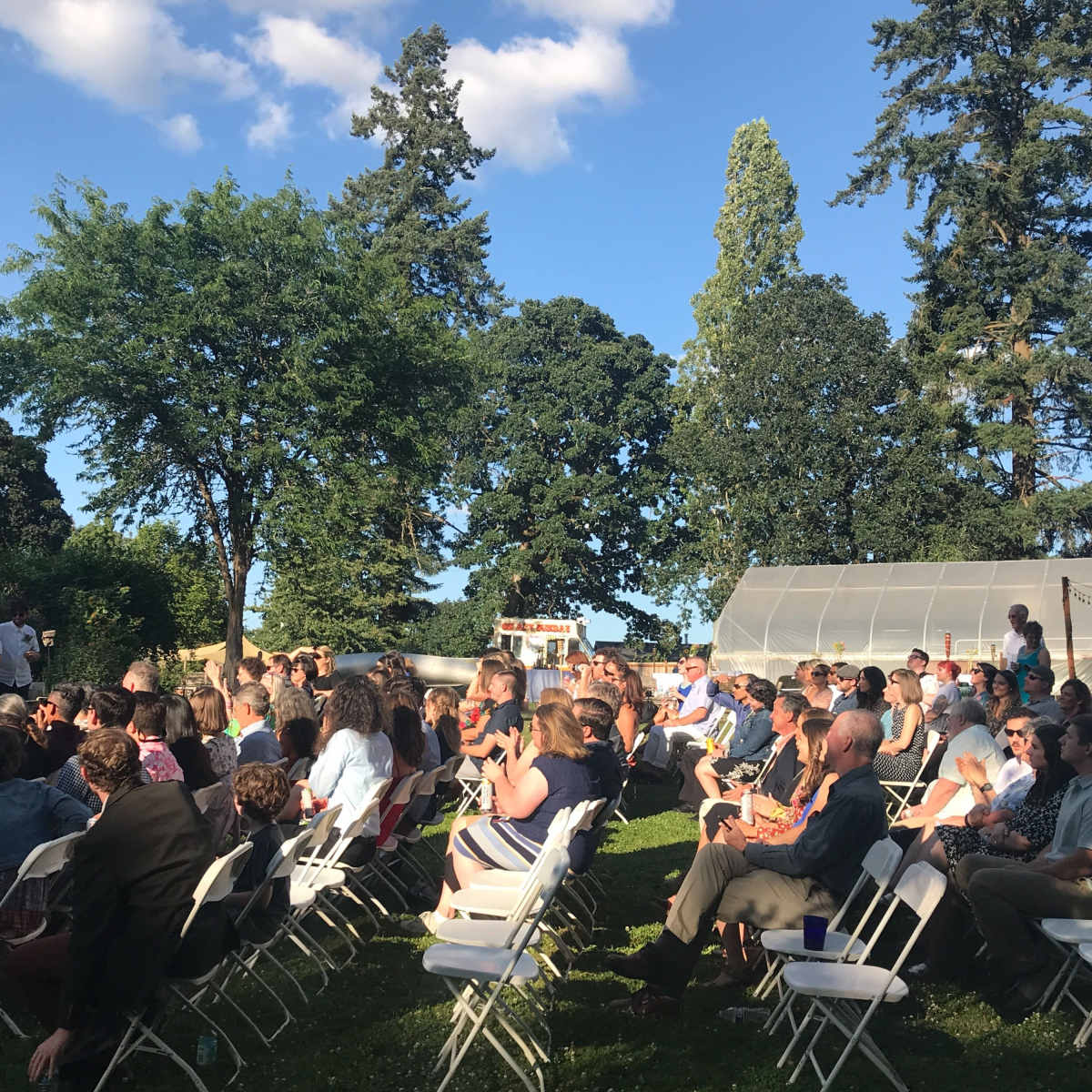 A large crowd of people sits in white folding chairs under trees during an outdoor event.