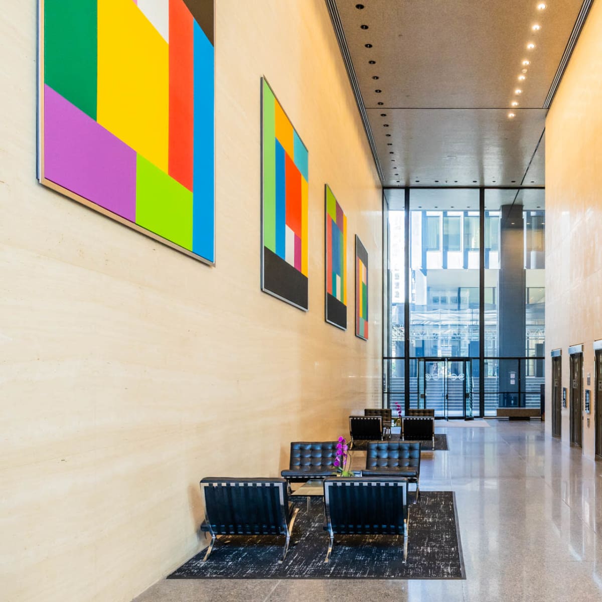 A modern lobby with a high ceiling, large windows, colorful abstract paintings on the walls, and an arrangement of seating near the elevators.