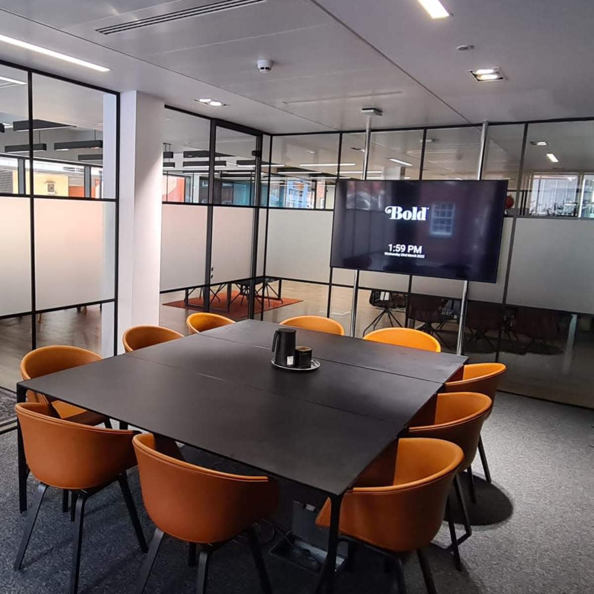 A modern meeting room with a black table surrounded by orange chairs, large glass walls, and a digital clock displaying the time.
