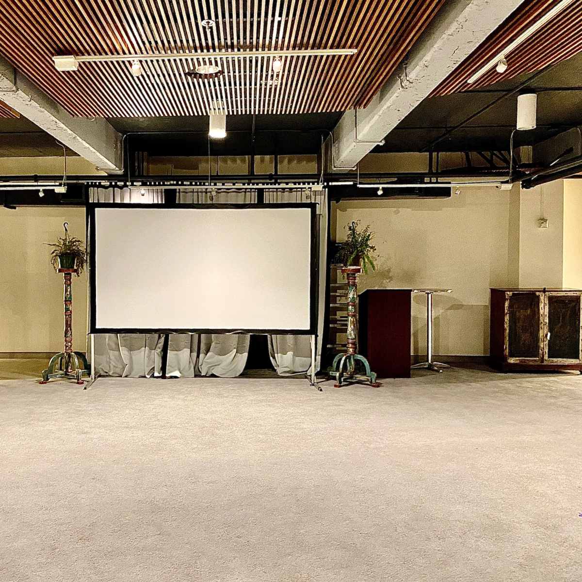 A spacious indoor venue featuring a large projection screen at the center, flanked by decorative plants and furniture.