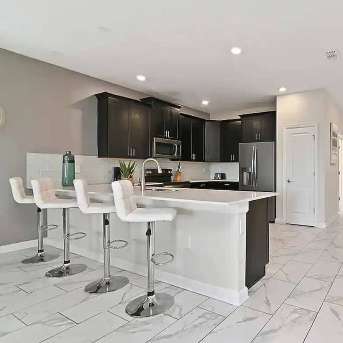 A modern kitchen area with black cabinets, a white island, and bar stools, leading into a hallway with light-colored walls and tile flooring.