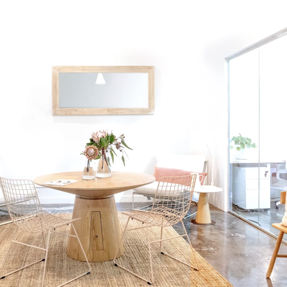 A bright, modern interior featuring a round wooden table surrounded by wire chairs, with potted plants and a large mirror on a white wall.