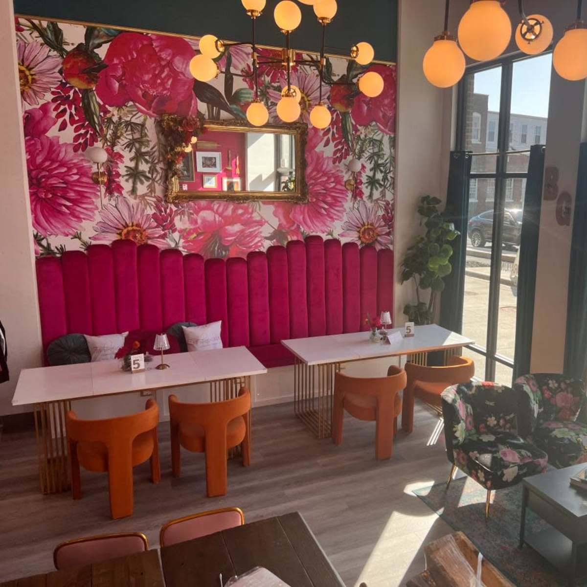 A modern café interior featuring a vibrant floral wallpaper, pink upholstered seating, and various tables with orange and floral-patterned chairs.