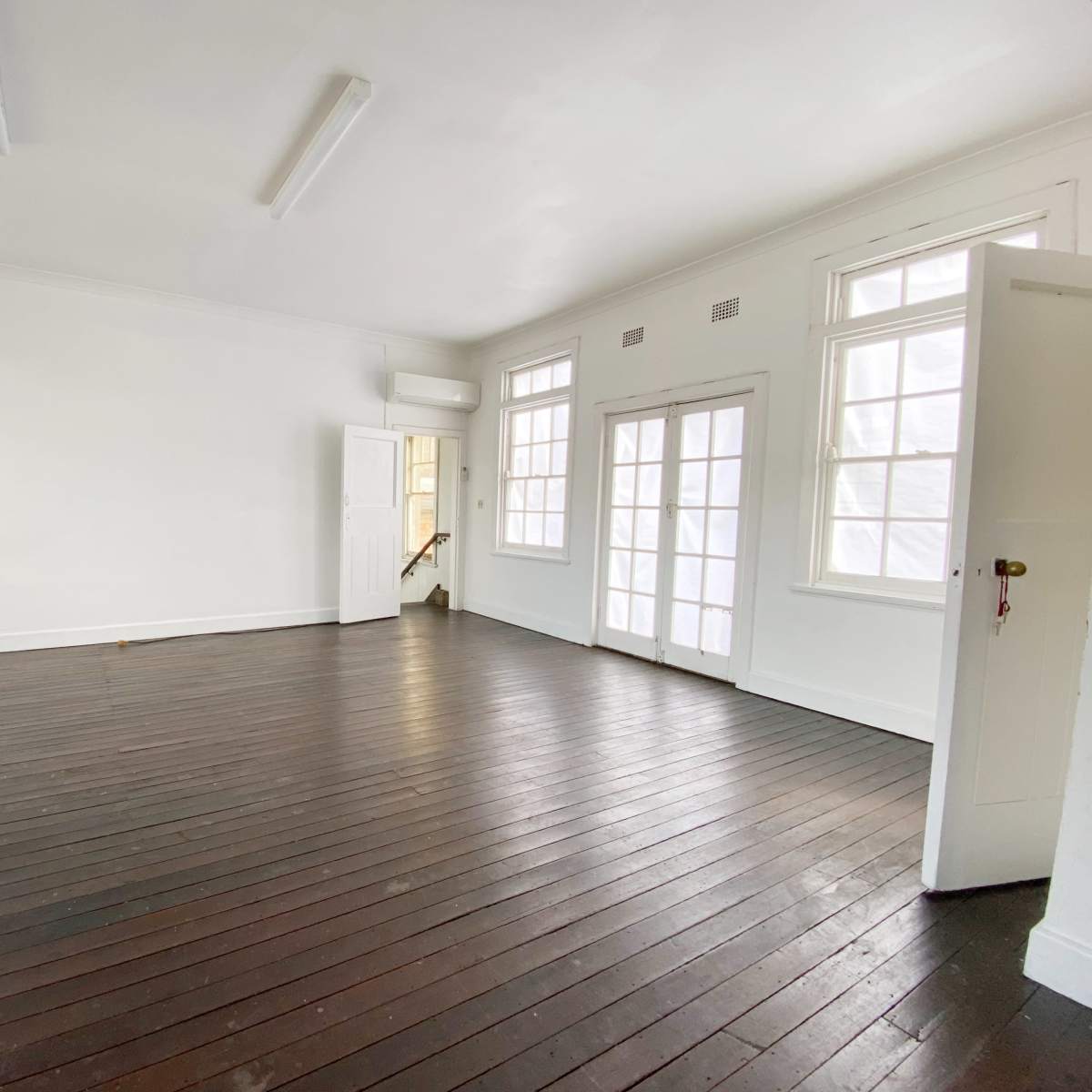 The room features wooden flooring, white walls, and large windows, with a door leading to another area.