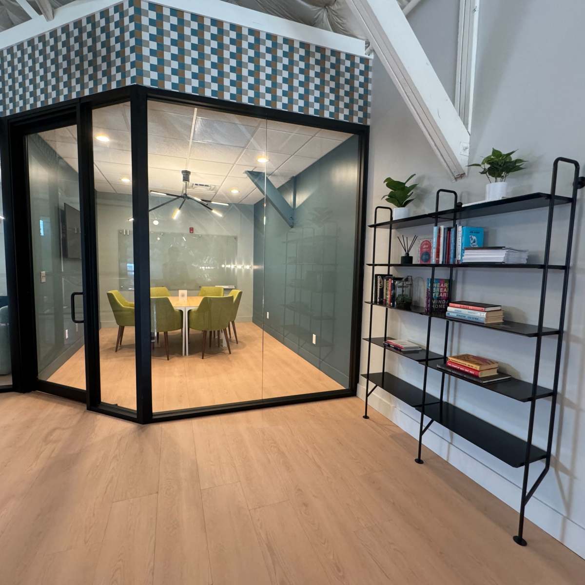 A modern office space features a glass-walled meeting room with a table and green chairs, alongside a black metal bookshelf filled with books and decorative items.