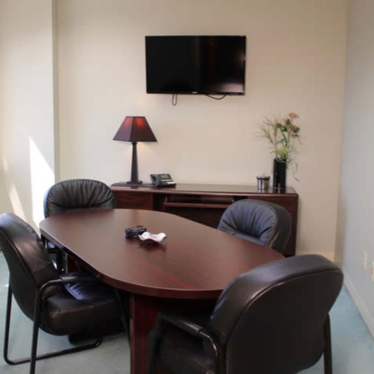 A small conference room with a wooden table, four black chairs, a wall-mounted television, and a lamp on a side table.