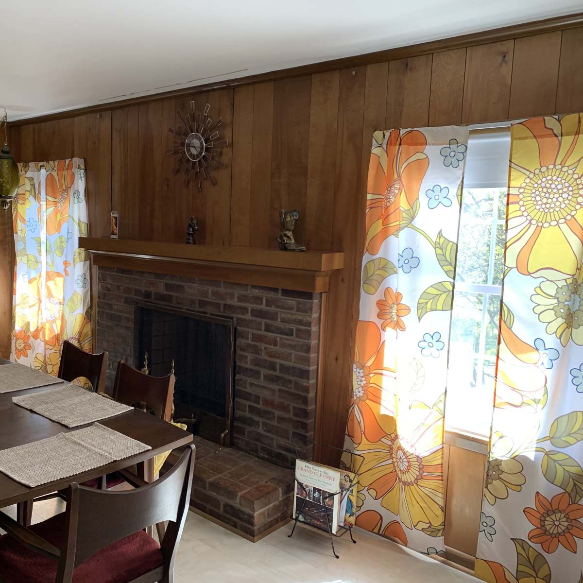 A dining area with a wooden table, chairs, a brick fireplace, and floral-patterned curtains on the windows.