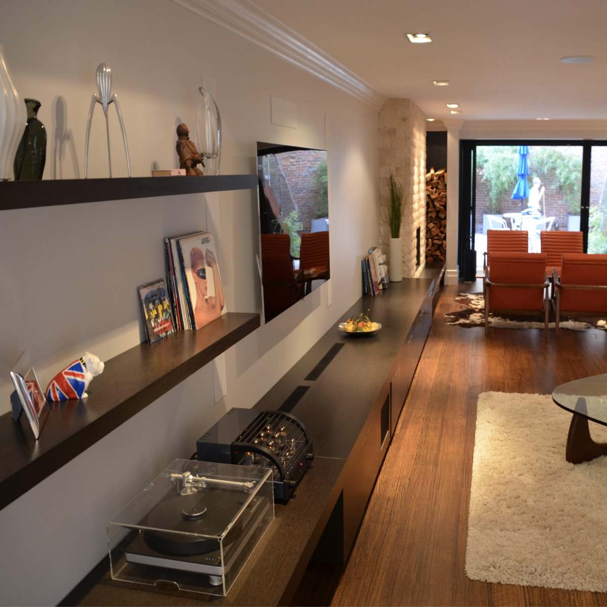 A modern living room featuring wooden flooring, a glass coffee table, and shelves displaying framed photos and decorative items.