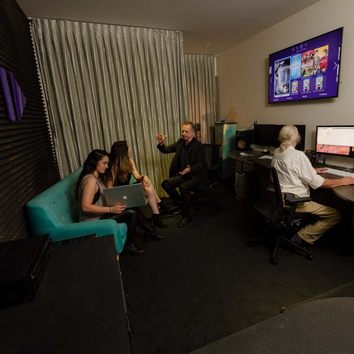 A group of four people is engaged in a discussion in a modern studio with computers and a large screen displaying digital content.