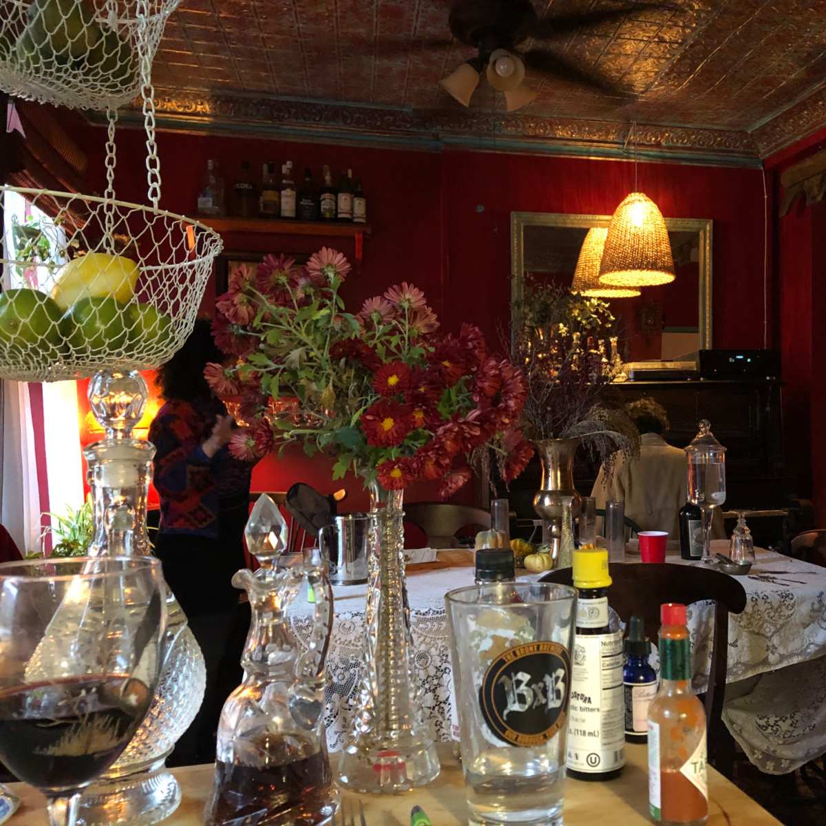 A cozy restaurant interior with a table set for dining, flowers in glass vases, and various bottles on display against a backdrop of red walls and vintage decor.