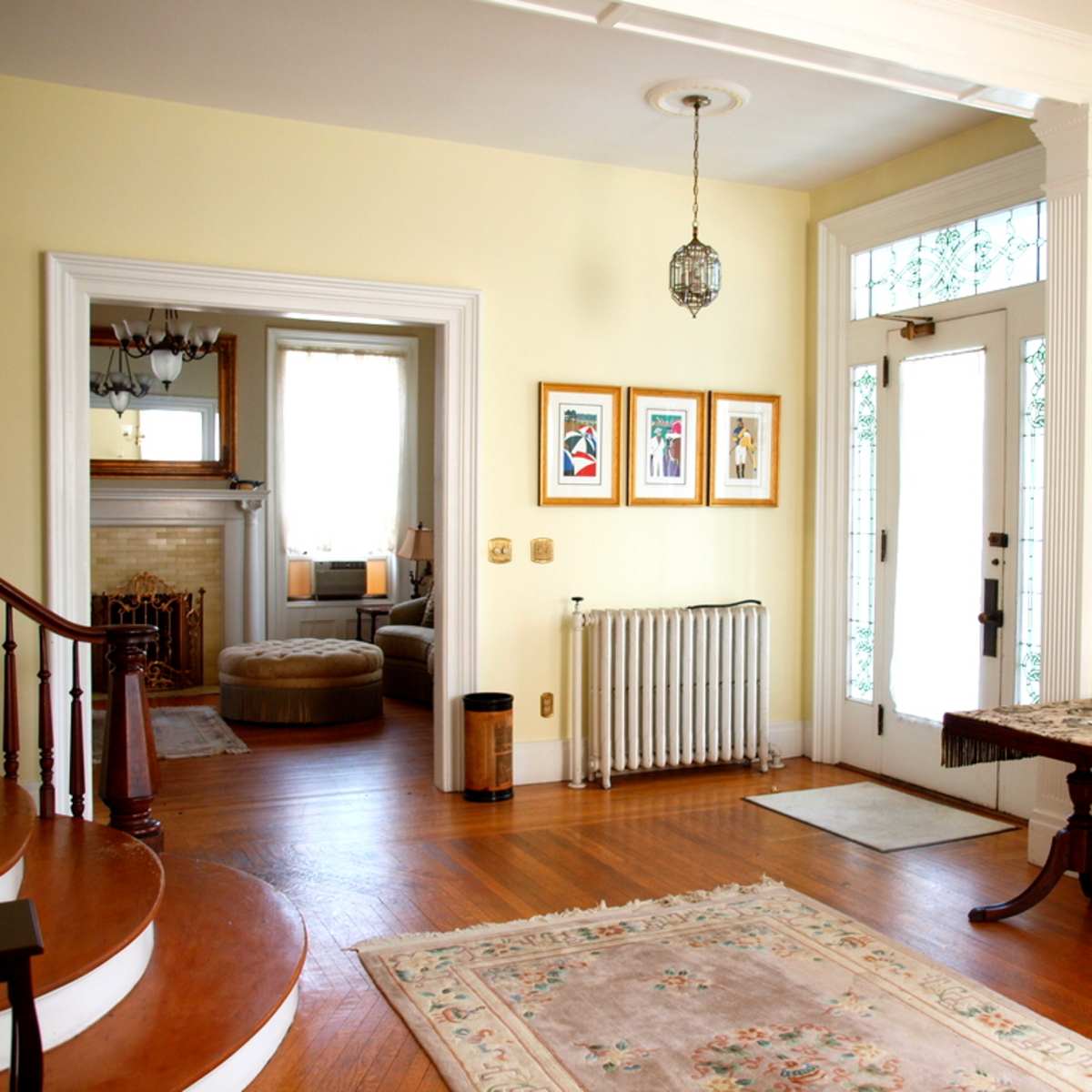 A bright entryway with a staircase, framed art on the wall, a radiator, and a door leading to an adjacent room.