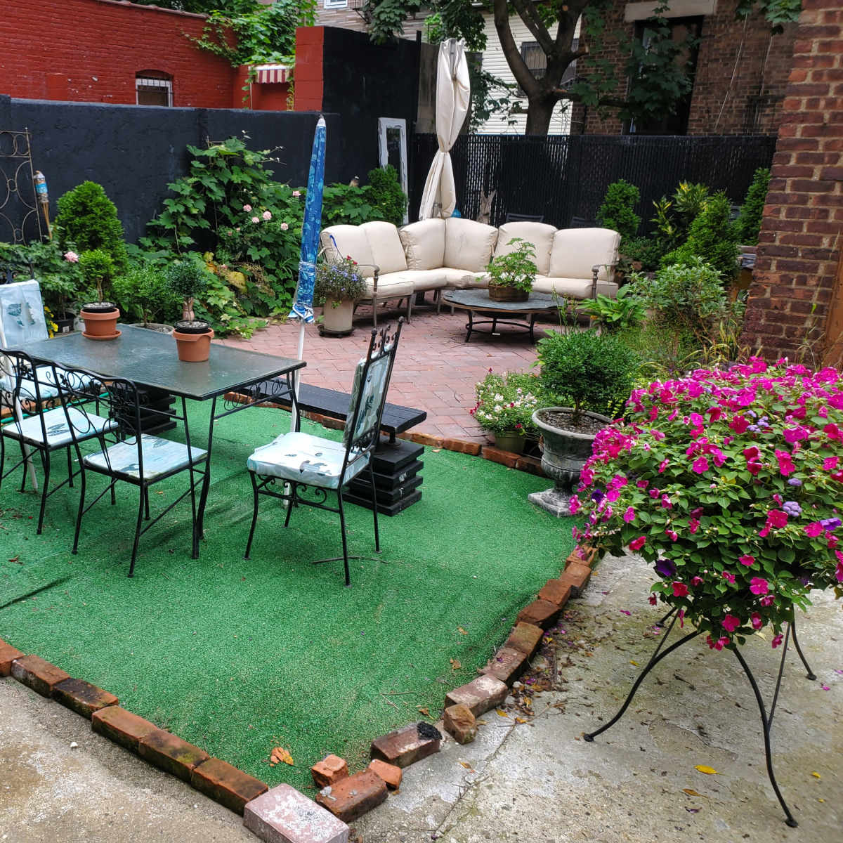 A backyard area with a green artificial grass surface, a dining table surrounded by chairs, and a seating arrangement with a sofa in the background, all framed by various plants and flowers.