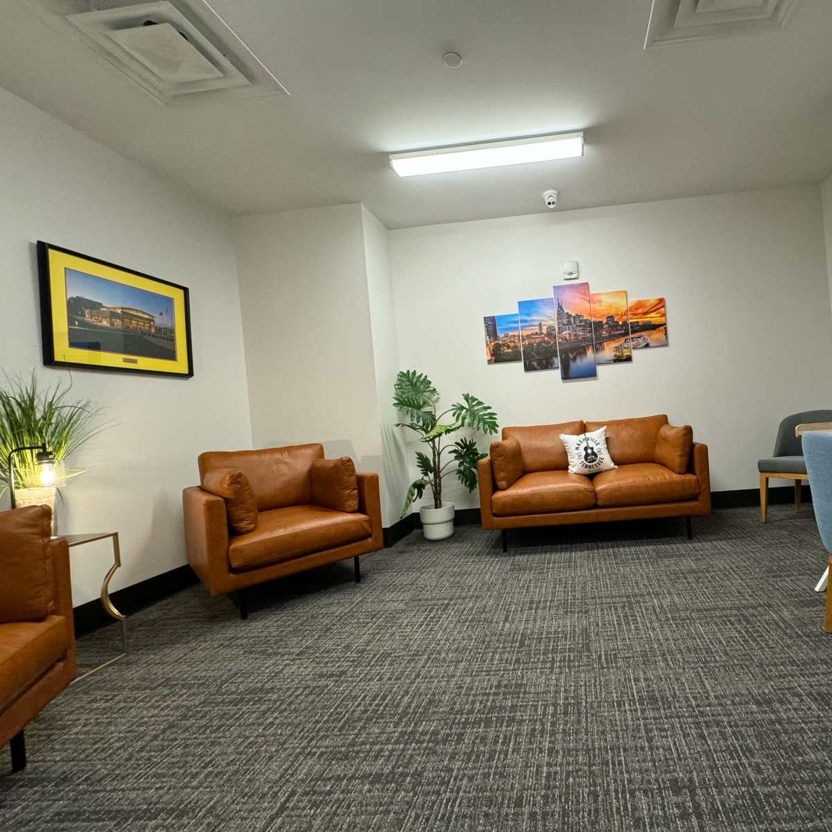 The room features two brown leather sofas, a blue chair, a desk, and decorative plants, with artwork on the walls.