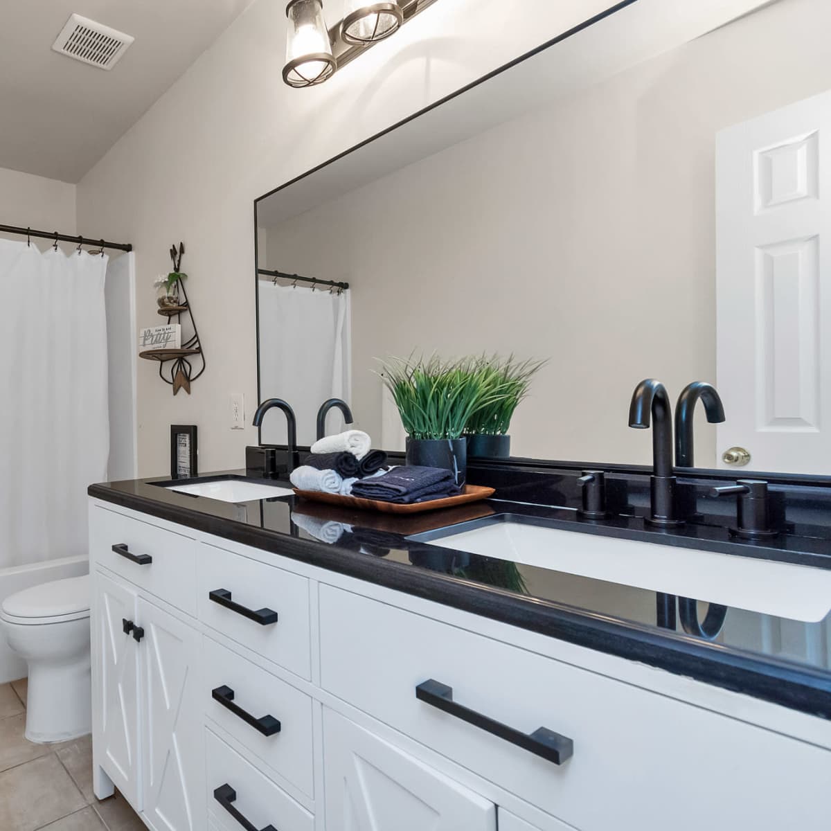 A modern bathroom features a white shower curtain, a black countertop with two sinks, and a large mirror above a white cabinet.