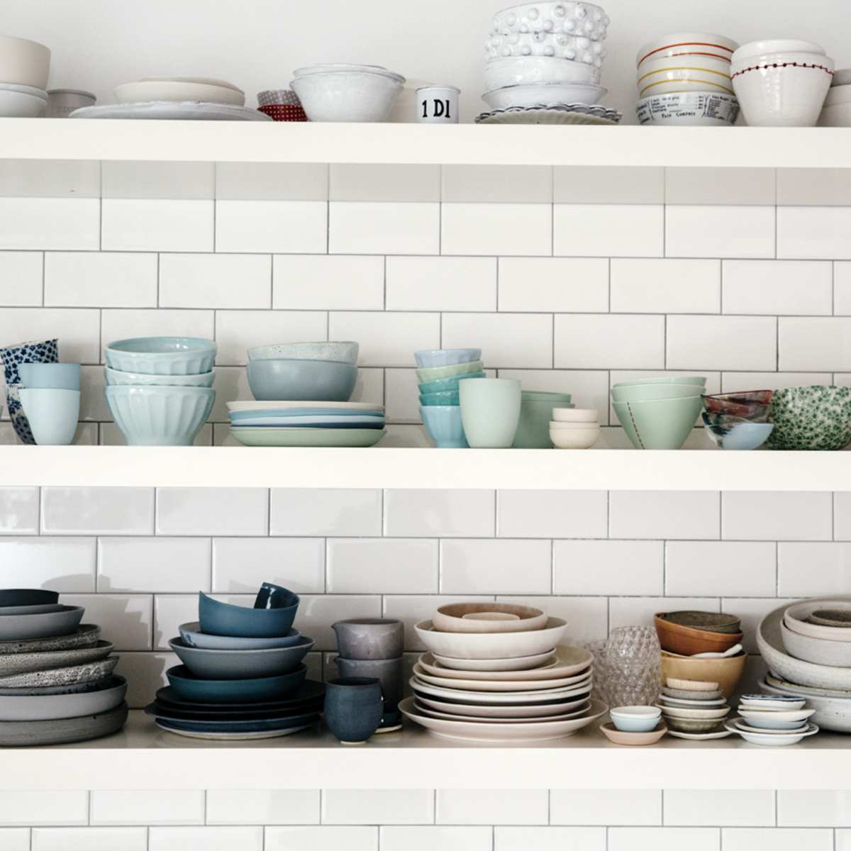 A collection of various plates, bowls, and cups is displayed neatly on two shelves against a tiled wall.