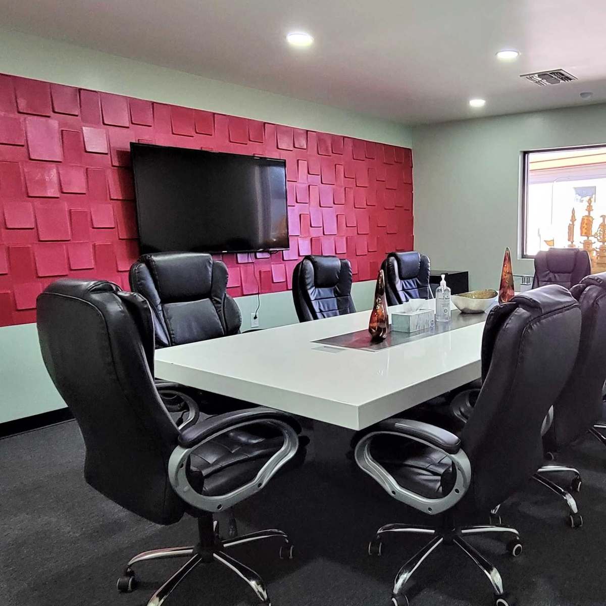 A modern conference room with a large white table surrounded by black ergonomic chairs, a television mounted on the wall, and a textured red accent wall.
