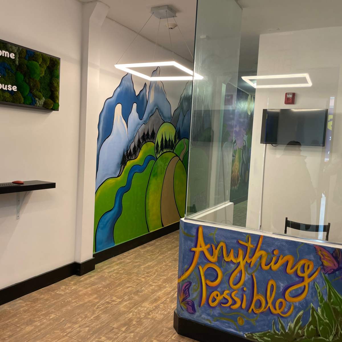 The interior of a community clubhouse features a welcome sign, a colorful mural of mountains and greenery, and a desk with a computer monitor.