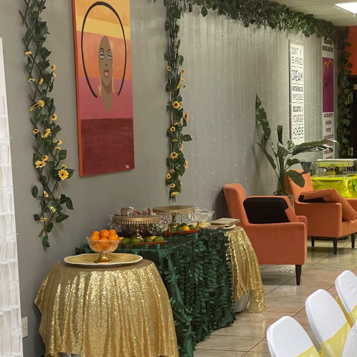 A decorated refreshment table covered with a golden tablecloth is set against a wall adorned with greenery and artwork.