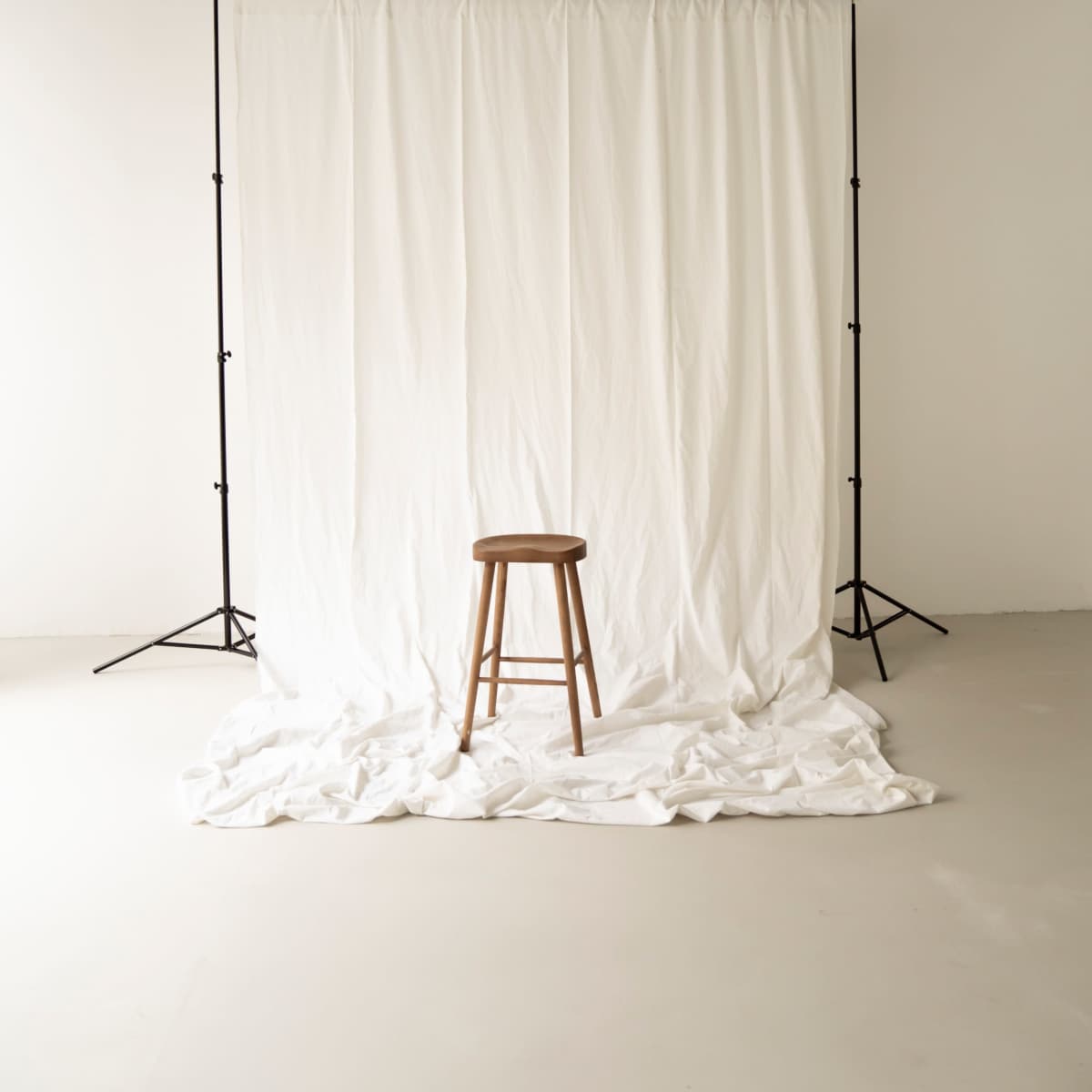 A wooden stool is positioned in front of a white backdrop draped on the floor.