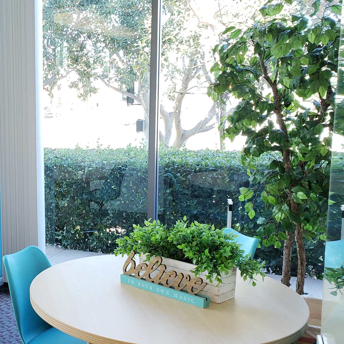 A bright meeting room with two round tables surrounded by blue chairs, decorated with potted plants and artwork on the walls.