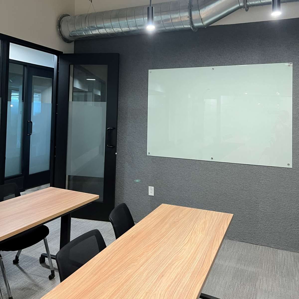 A modern conference room with two wooden tables, black chairs, and a glass whiteboard on a gray wall.