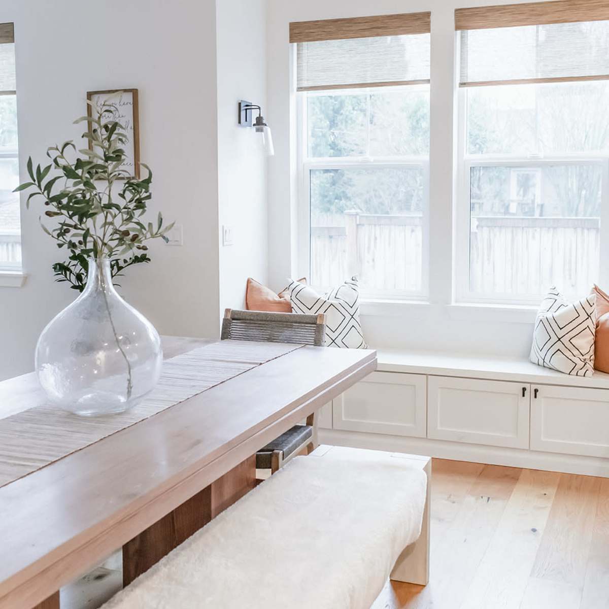 A dining area features a long wooden table with a bench, a large glass vase with foliage, and bay windows with cushions.