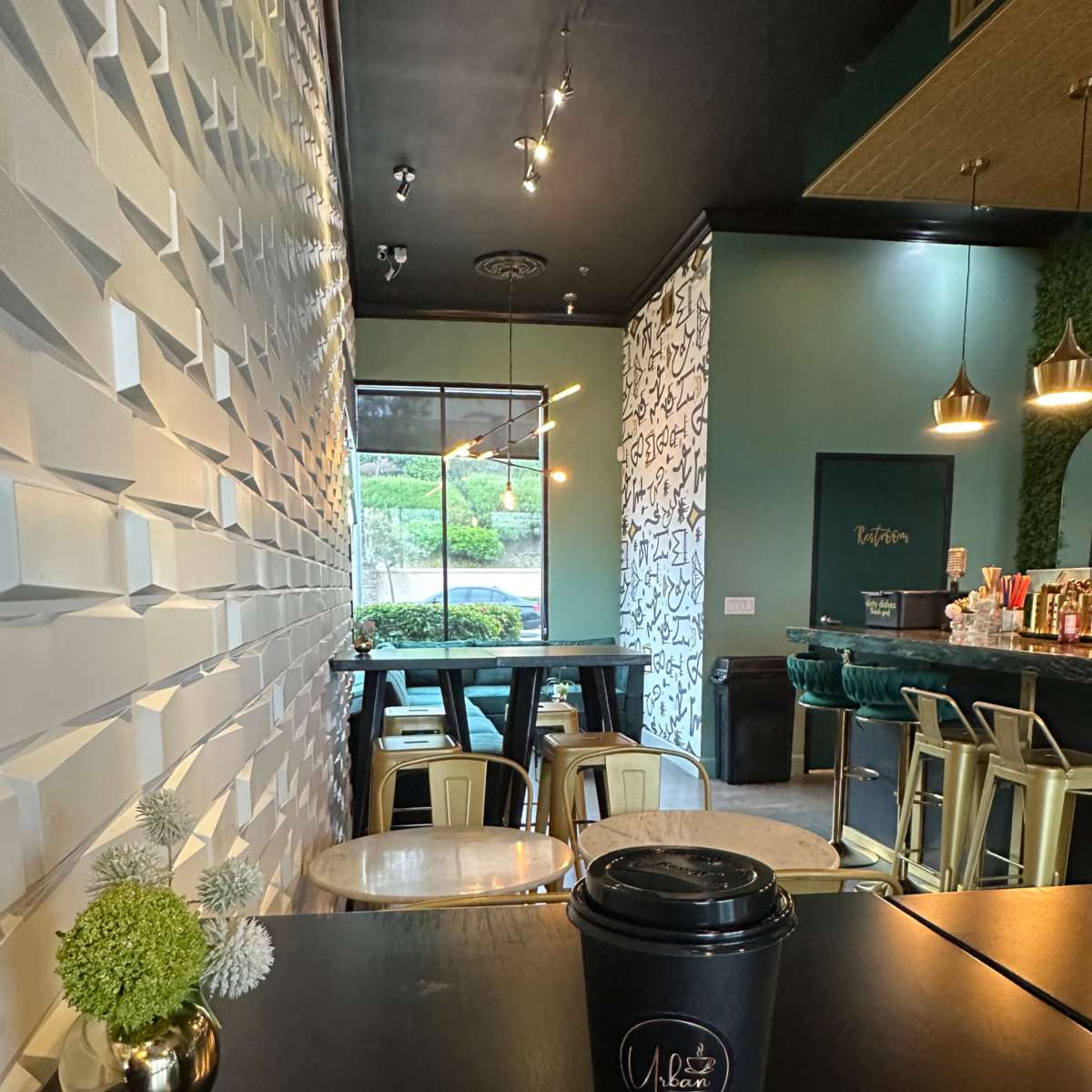 A modern cafe interior with a decorative wall, dark tables, and a takeaway coffee cup prominently displayed in the foreground.