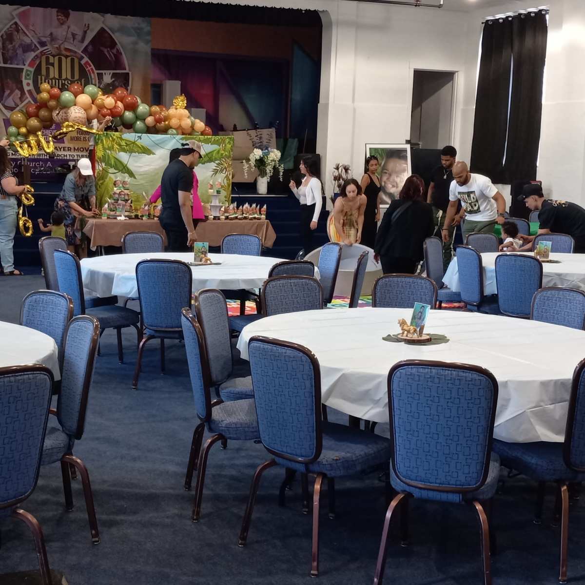 A large event space with several round tables covered in white tablecloths and green overlays, where people are arranging decorations and preparing for an event.