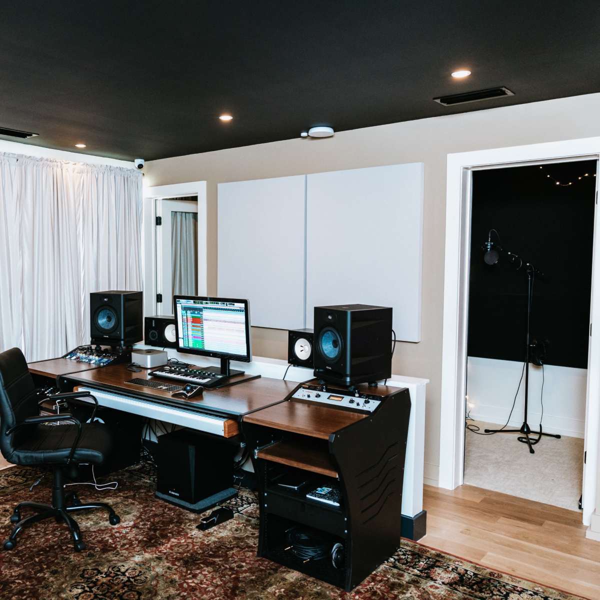 A music production studio featuring a desk with computer monitors, studio speakers, and a keyboard, along with soundproof panels and a microphone setup in the recording area.