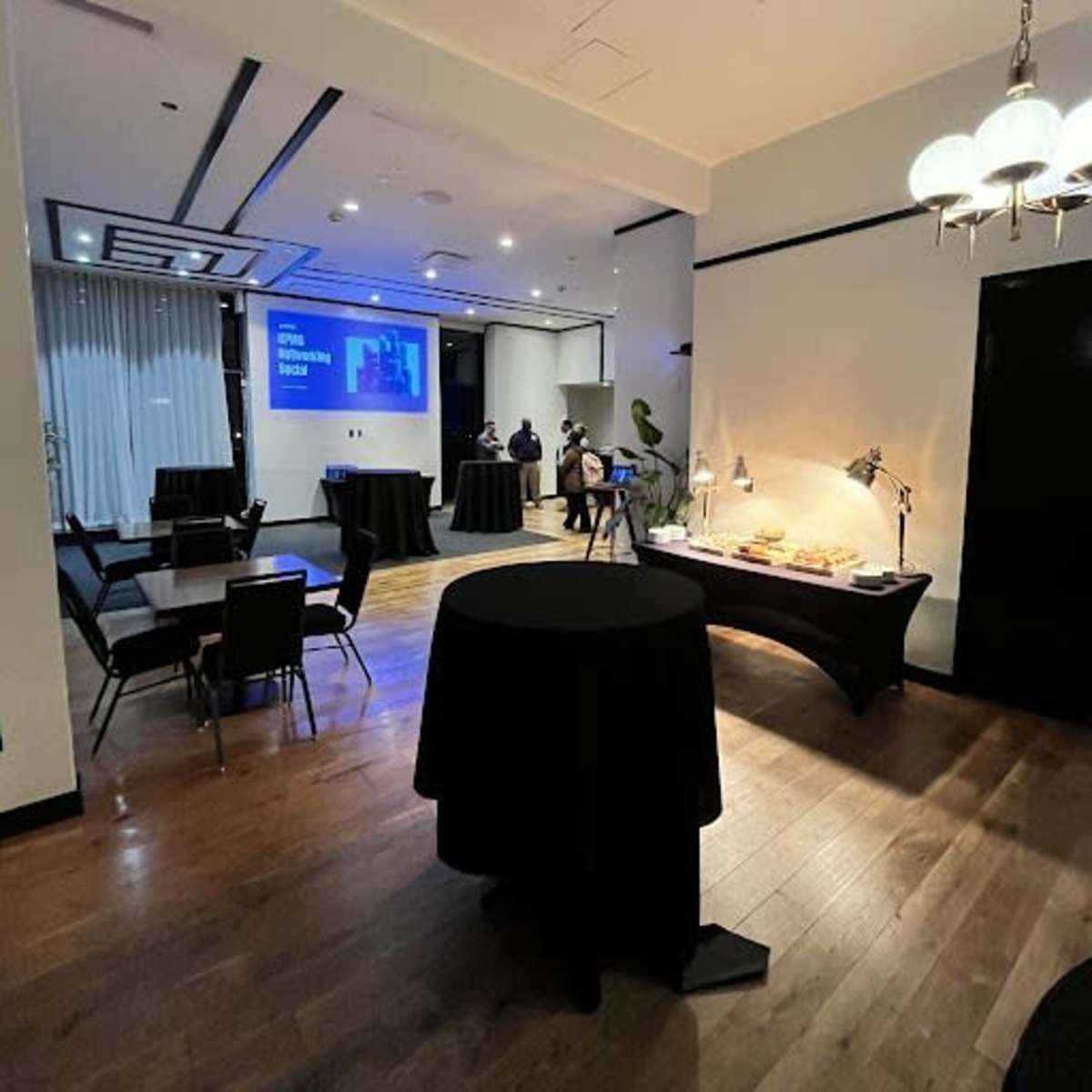 A spacious event room with black tables, a screen displaying a presentation, and a buffet setup against the wall.