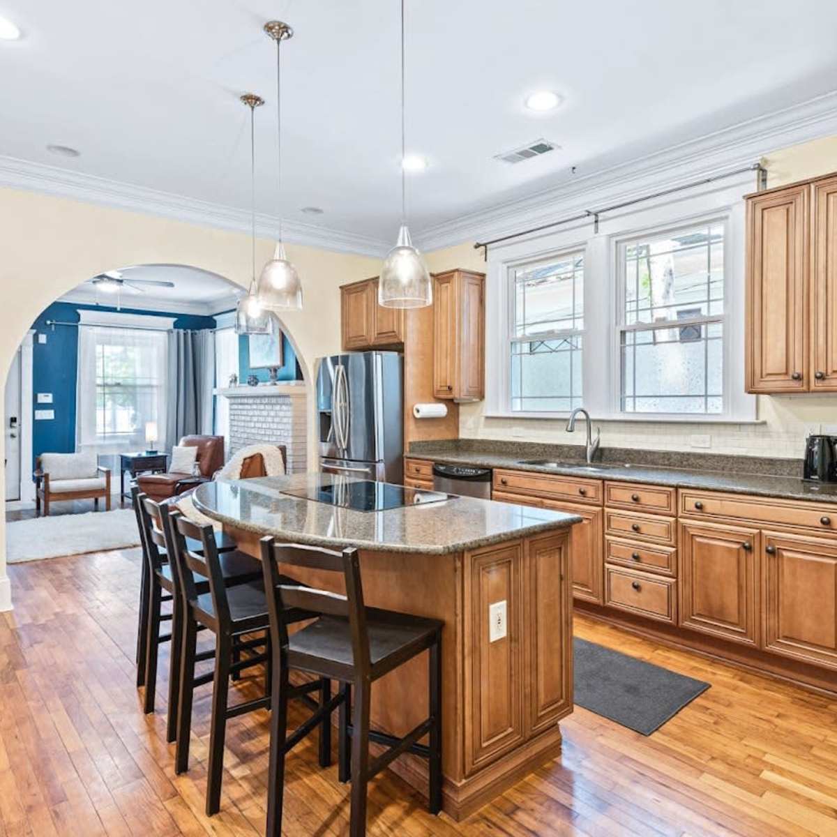 A modern kitchen with wooden cabinets, a central island with seating, and an adjoining living area visible through an archway.