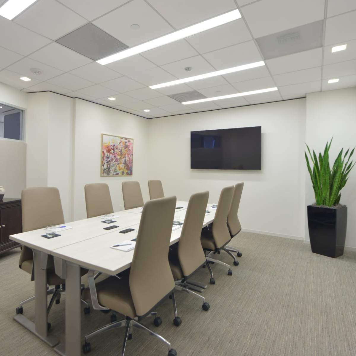 A modern conference room features a long rectangular table surrounded by beige chairs, with a wall-mounted TV and a large plant in the corner.