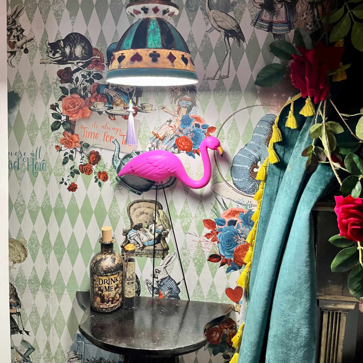 A colorful corner with a vintage lamp, a pink flamingo figure, a decorative bottle labeled "DRINK ME," and a patterned wallpaper adorned with flowers and animals.