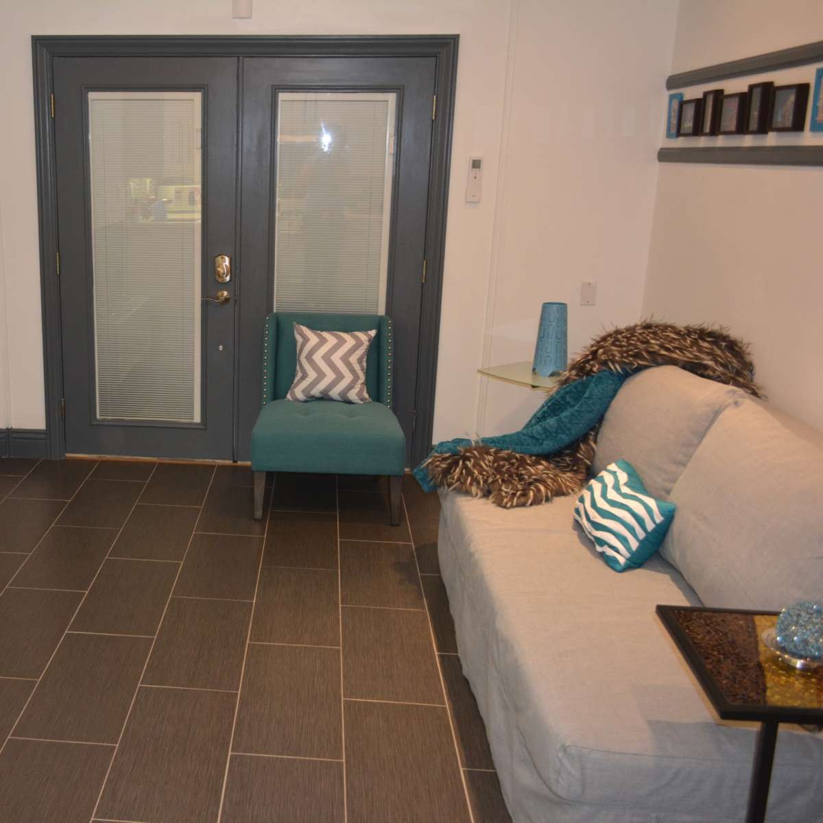 A modern living room features a gray sofa, a turquoise chair, a television on a white stand, and a glass table with decorative items.