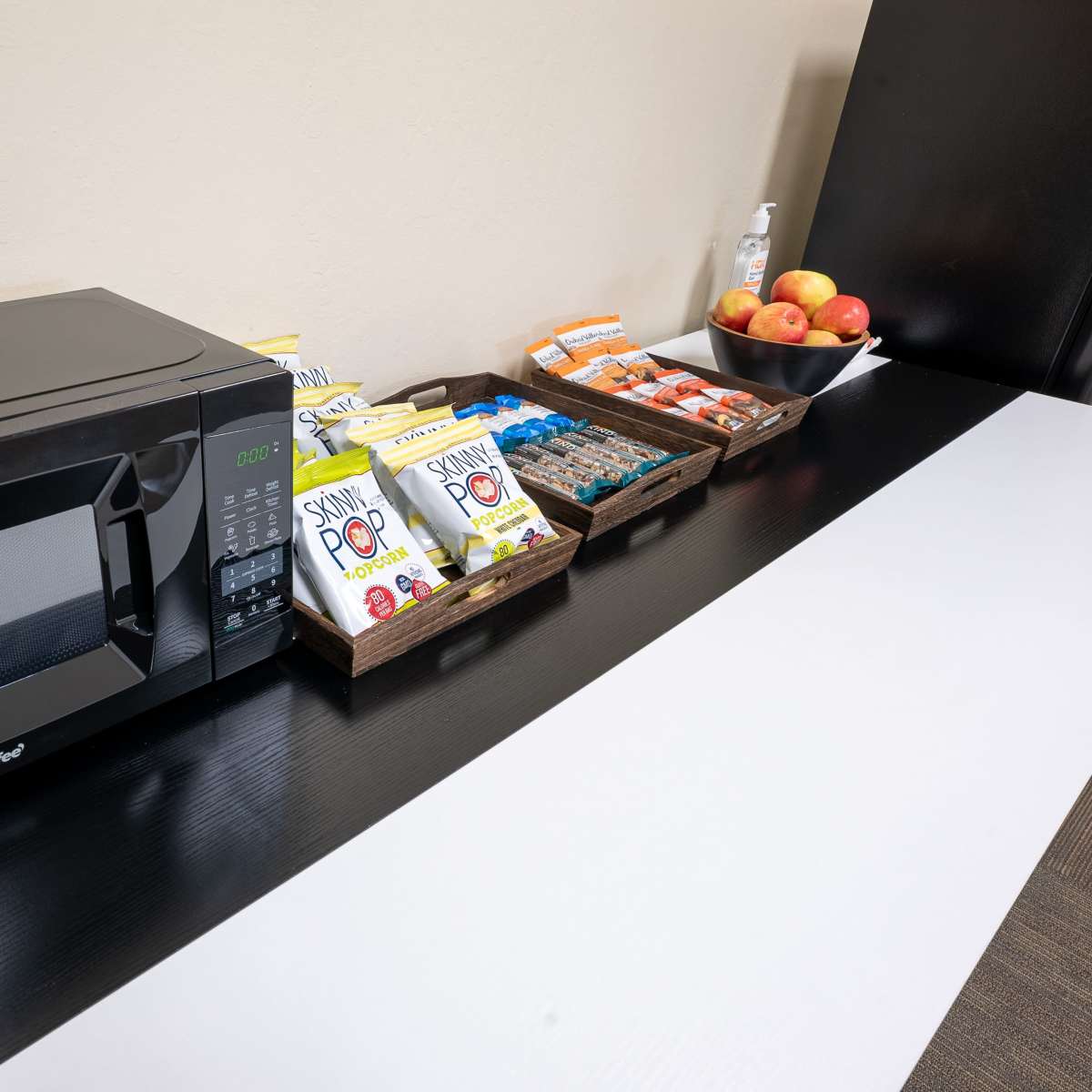 A black microwave is placed on a countertop next to various snacks and apples arranged in baskets.